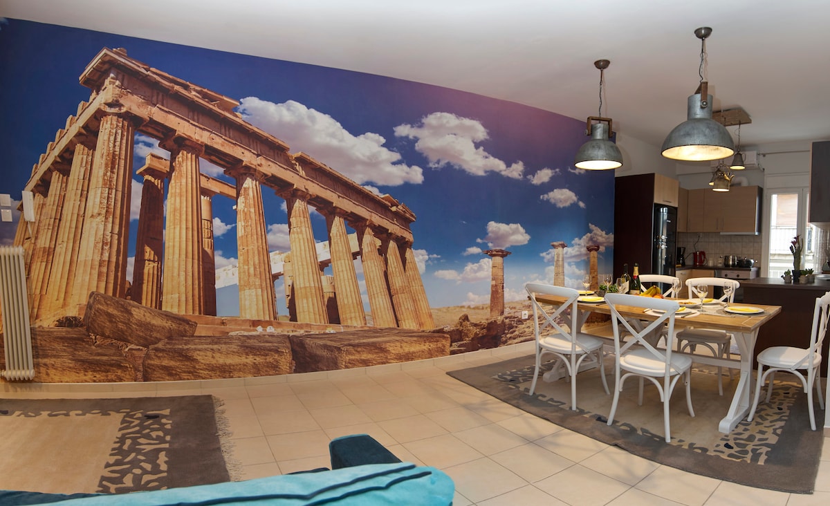 A spacious living and dining area features a large mural of the Parthenon, providing a striking backdrop. A wooden dining table with white chairs is set for meals, while the room is illuminated by overhead pendant lights. Neutral tones and natural light enhance the ambiance.
