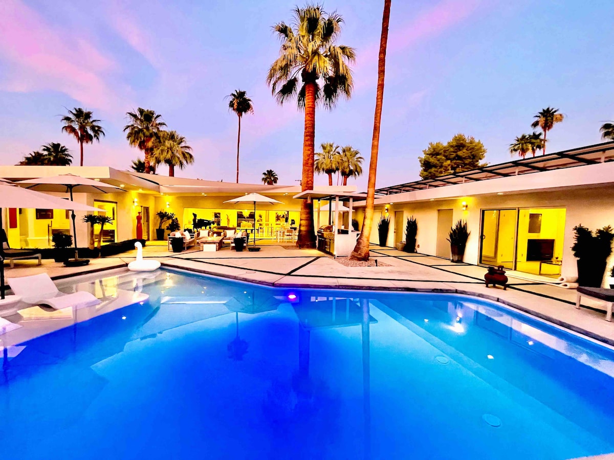 An inviting pool area is surrounded by palm trees, with a shimmering blue surface reflecting the evening sky. Sun loungers and umbrellas are positioned nearby, alongside spacious outdoor seating areas. Soft lighting enhances the modern architecture of the private compound in a tranquil setting.