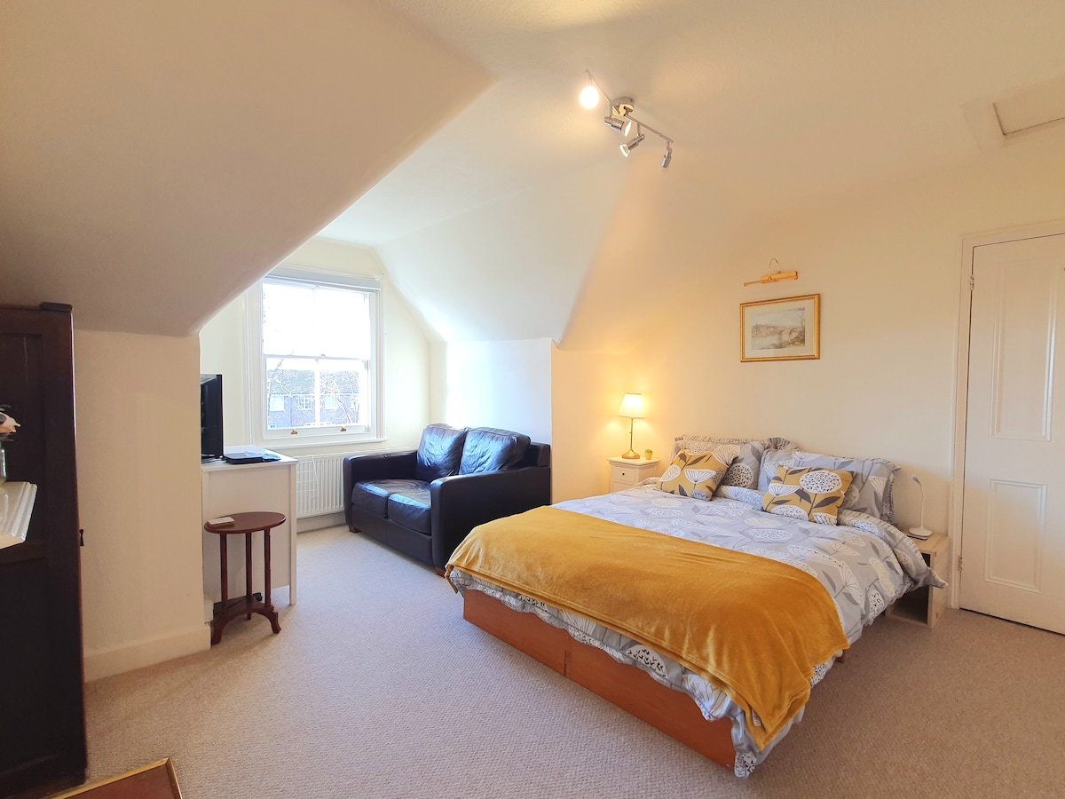 A spacious king-size bedroom features a large window allowing natural light to fill the space. A comfortable brown leather armchair sits adjacent to the bed, which is layered with decorative bedding. A small table and television are positioned against the wall, enhancing the room's functionality.