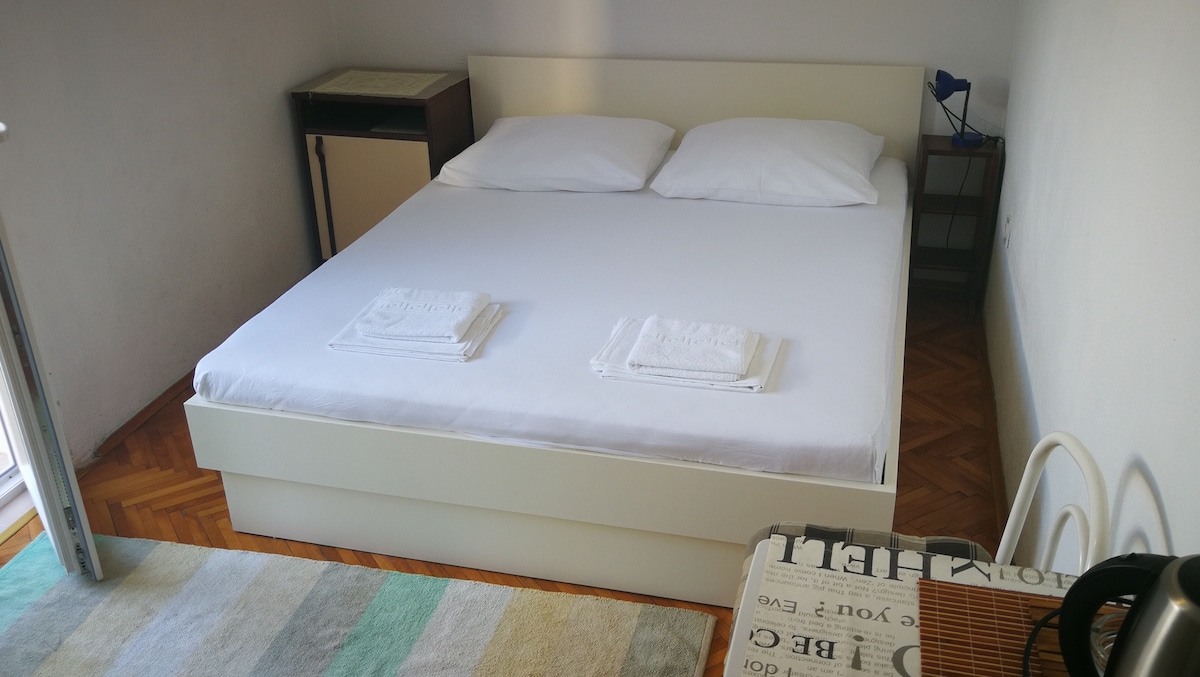 A well-lit sleeping area features a spacious bed with crisp white linens and two neatly folded towels on top. A small table is positioned beside the bed, along with a chair. The room's floor is covered in a striped rug, enhancing the layout.