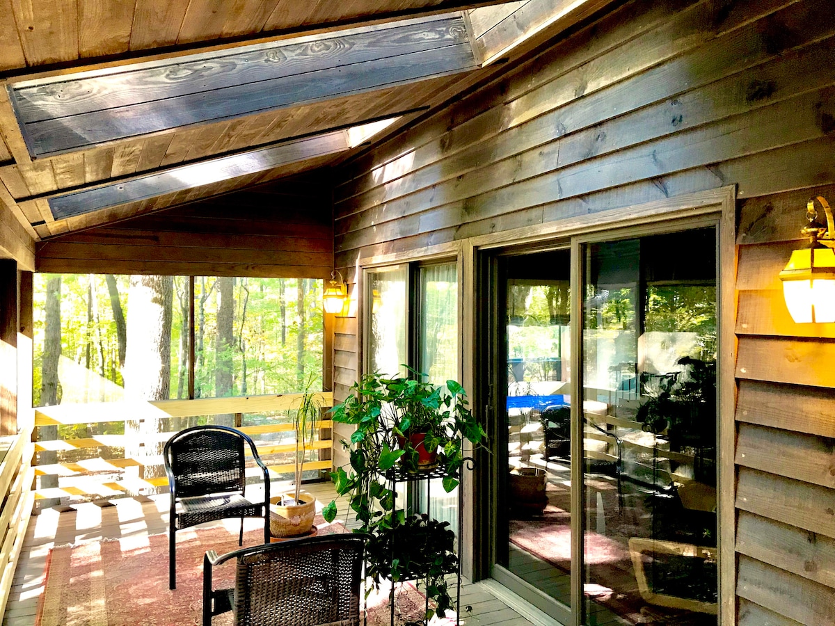 A wraparound porch is featured with large glass doors, providing a view of the surrounding forest. Two black chairs and a potted plant are arranged on a wooden deck, while natural light filters in through a slanted roof.