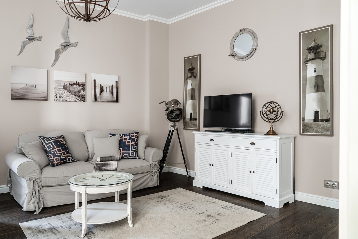 The living area features a light-colored sofa adorned with decorative cushions, a round glass coffee table, and a stylish TV cabinet. Two framed images of lighthouses are displayed on the walls, while a vintage projector stands beside the TV, enhancing the coastal theme.