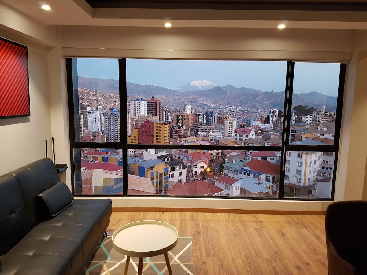 A panoramic city view is showcased through a large window, revealing a landscape of urban buildings against a backdrop of mountains. A comfortable sofa and a round coffee table are positioned in front, allowing for a relaxing spot to enjoy the scenery.