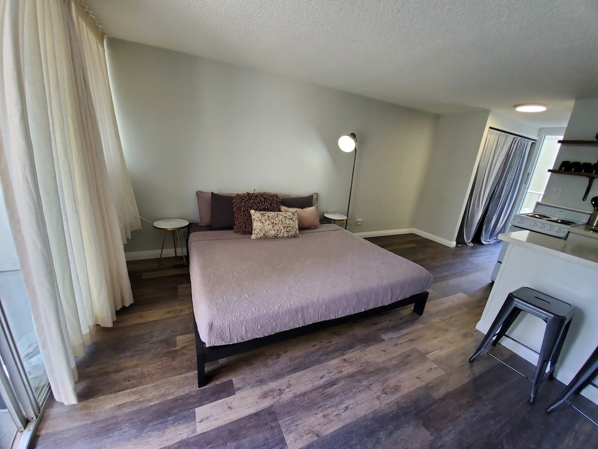 A spacious sleeping area is furnished with a large bed topped with neutral bedding and decorative pillows. Natural light filters through sheer curtains, highlighting the warm wood flooring. A modern floor lamp stands beside the bed, and a small side table offers additional space.