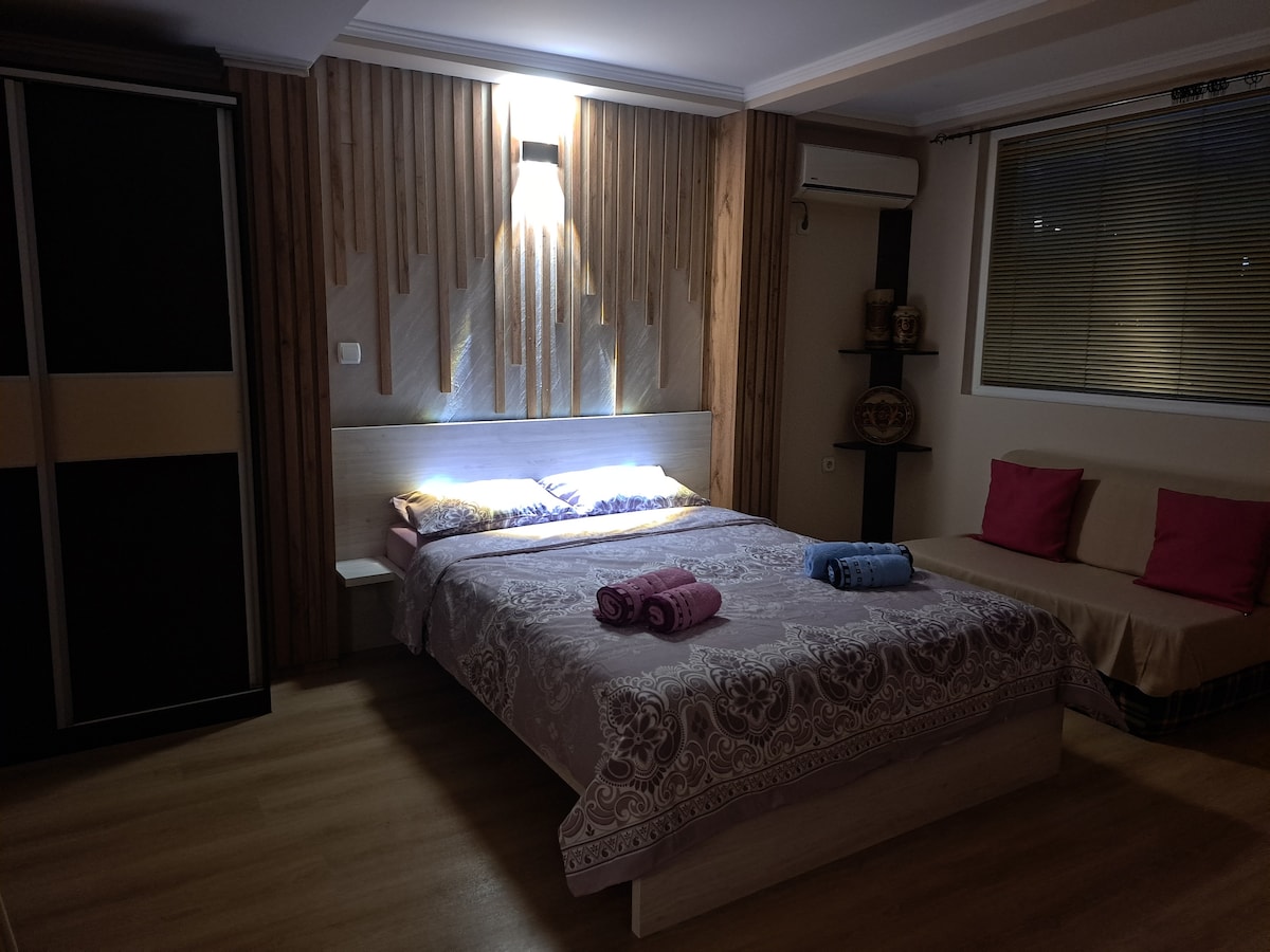 A cozy bedroom is presented with a spacious bed featuring a decorative coverlet. Soft pillows are arranged on the bed alongside neatly rolled towels in shades of pink and blue. A seating area is included, complemented by warm wooden accents and ambient lighting.