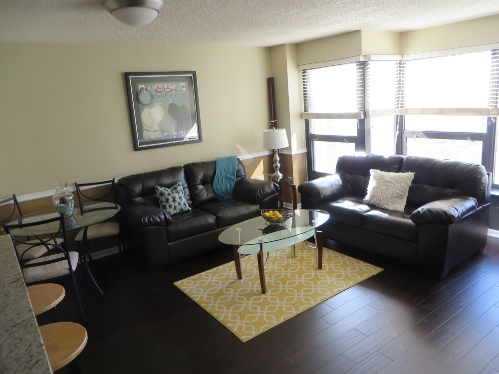 The living area features two leather sofas arranged around a glass coffee table, complemented by a decorative area rug. A round dining table is set for four, while large windows provide ample natural light, framed by stylish blinds.