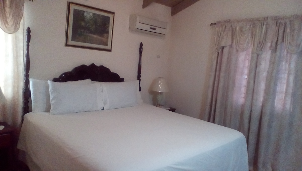 A spacious bedroom features a large bed dressed in white linens. Soft light filters through sheer curtains, illuminating the room. An air conditioning unit is mounted above, and a lamp sits on a nightstand beside the bed. A framed painting adds a touch of decor on the wall.