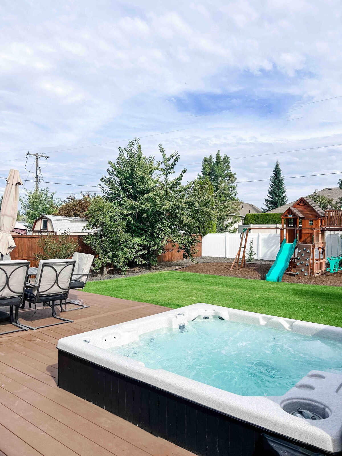 A spacious backyard is shown featuring a hot tub bordered by comfortable chairs. A playground with a slide and climbing structure is visible, alongside lush green grass and trees, creating a setting for relaxation and recreation.