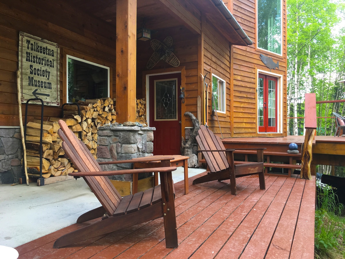 A welcoming outdoor space features two wooden adirondack chairs on a red deck, positioned near a stone entryway. Stacked firewood is visible to the side, complemented by a decorative sign. The entrance showcases large windows and a bright red door.