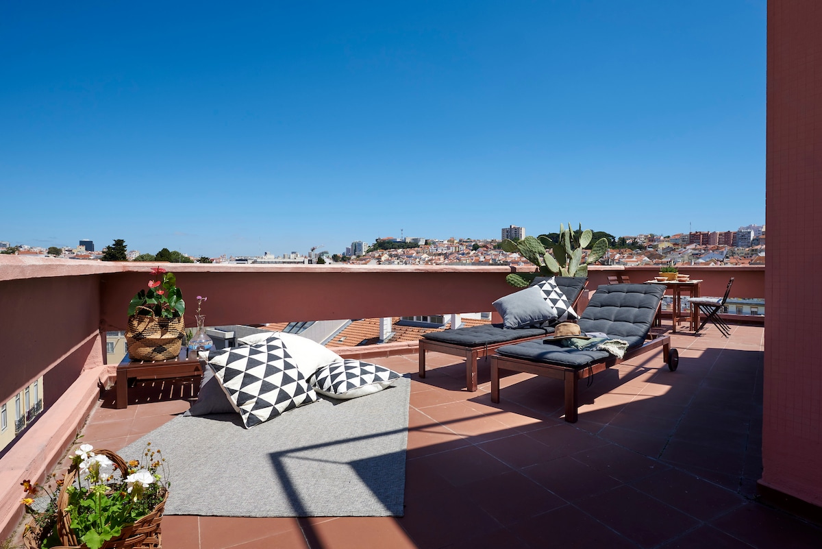A private terrace features comfortable seating arrangements, including lounge chairs and cushions on a textured rug. Potted plants add greenery, while expansive views of the city skyline are visible against a clear blue sky.