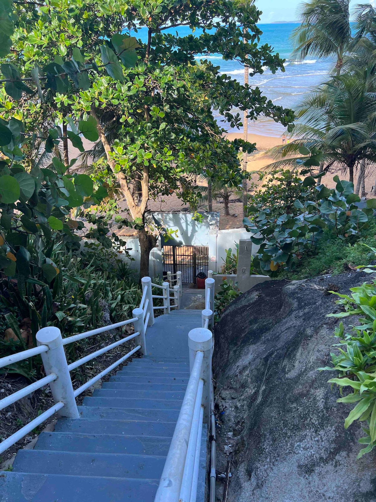 A set of concrete steps leads down towards a sandy beach, surrounded by lush greenery. Palm trees are visible in the background, and a gate is seen at the base of the steps, providing access to the beachfront.