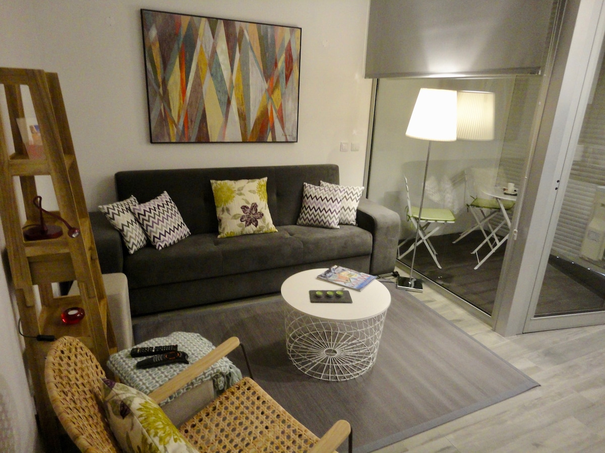 The living room features a comfortable sofa adorned with decorative pillows, complemented by a round coffee table in a light color. A chair made of natural materials adds texture, while a colorful wall art piece enhances the space. Light from an adjacent balcony illuminates the room.