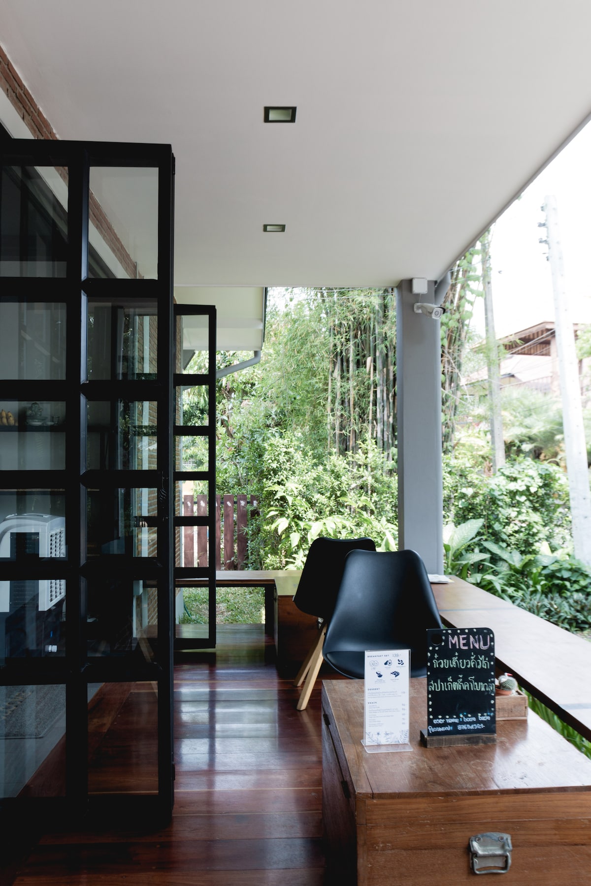 A serene outdoor seating area is highlighted, featuring wooden benches and sleek black chairs. Lush greenery surrounds the space, creating a calming environment. A small menu board and informational signs are displayed nearby, inviting guests to explore dining options.