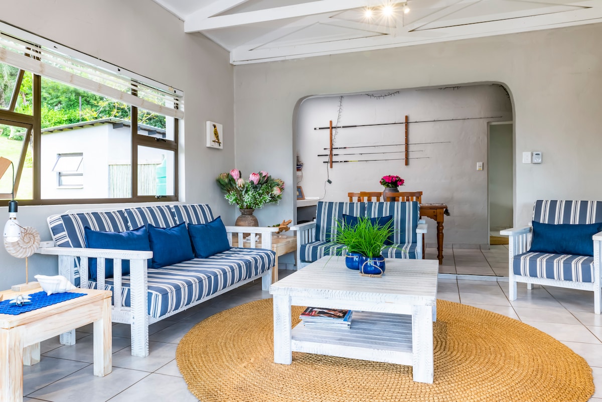A spacious living area features striped sofas with blue pillows, arranged around a light-colored coffee table. A natural fiber rug lies beneath, while large windows let in ample light. A dining area is visible in the background, accented by floral arrangements and minimal decor.