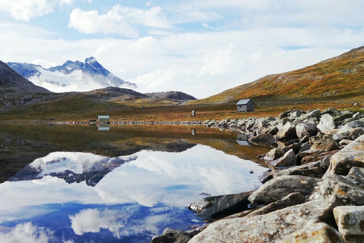 A serene landscape is depicted, featuring a calm body of water reflecting the sky and surrounding mountains. Rocky shorelines are visible in the foreground, while two small, simple structures stand in the distance against a backdrop of grassy hills and peaks.