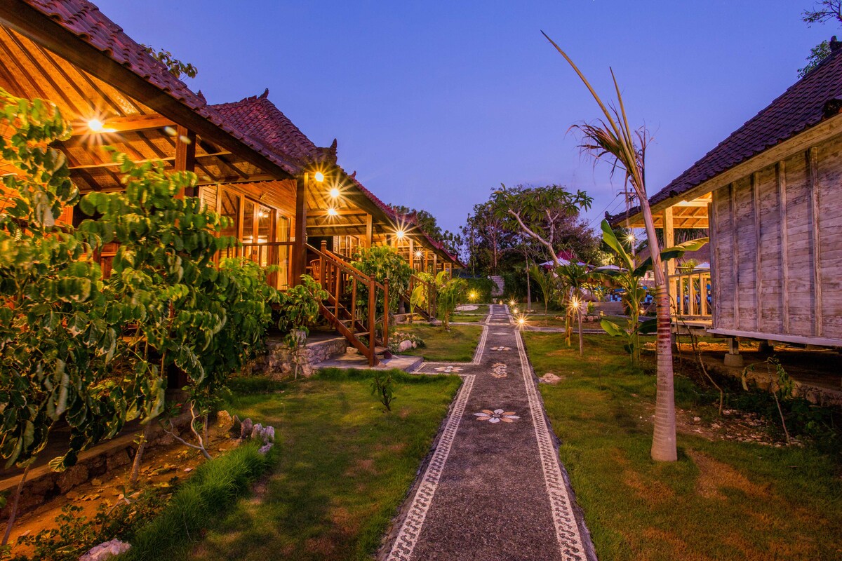 Wooden cottages featuring a blend of traditional and modern design are illuminated by soft lighting in the evening. A stone pathway, bordered by lush greenery, leads through a well-maintained garden setting. Tropical plants and landscaping enhance the natural ambiance of the surroundings.