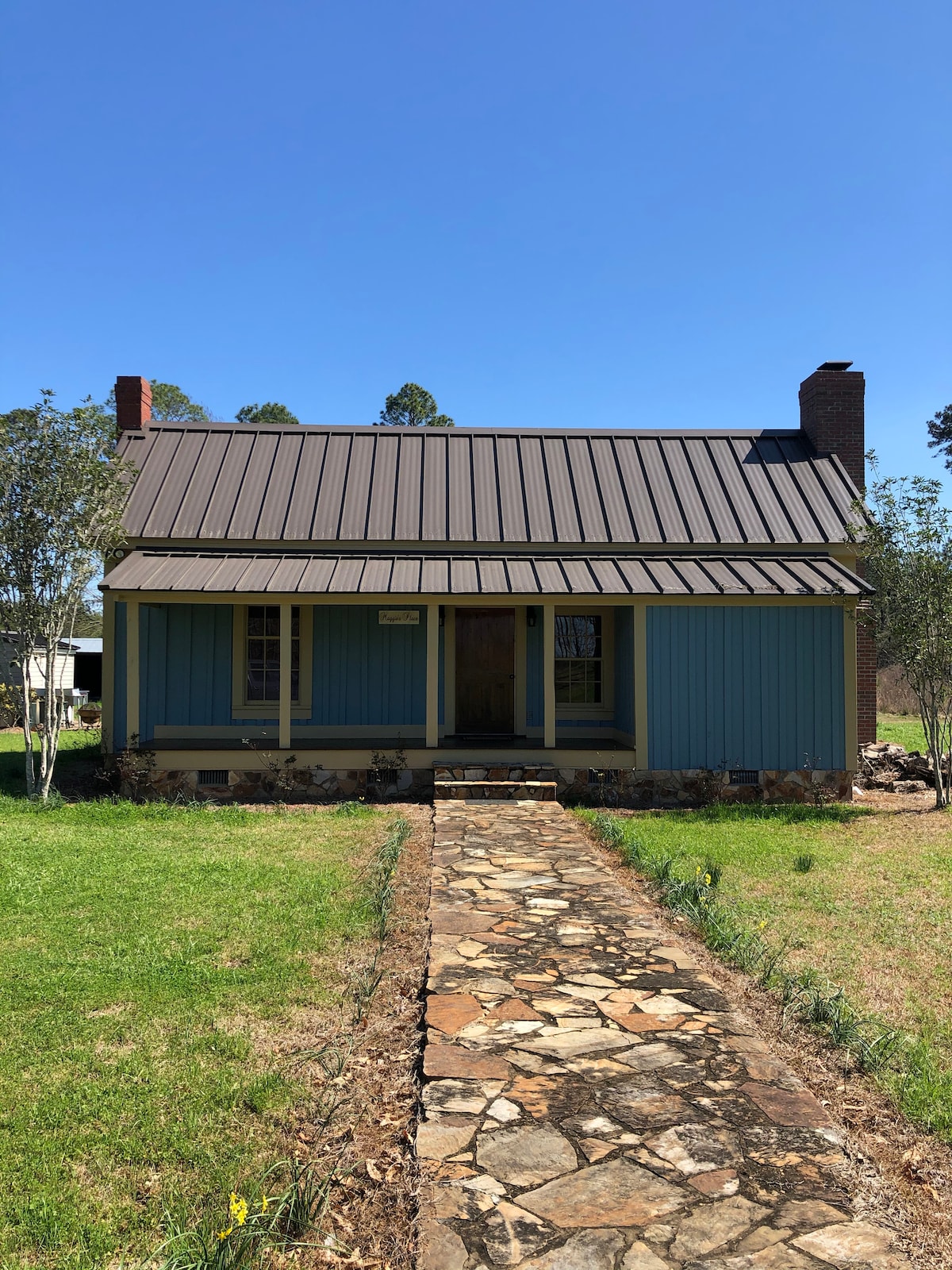 A quaint cabin features a blue exterior with wooden accents. The front porch is framed by two small trees, and a stone walkway leads to the entrance. A metal roof completes the charming countryside aesthetic under a clear blue sky.