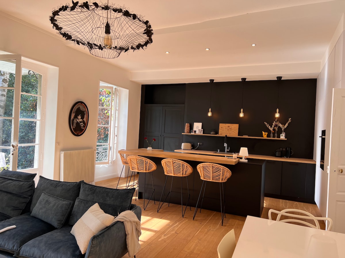 A modern kitchen and living area features a spacious countertop with bar seating. Two large windows provide ample natural light, and a unique chandelier hangs from the ceiling. A cozy sofa and dining table complete the open-concept space, offering a welcoming environment.