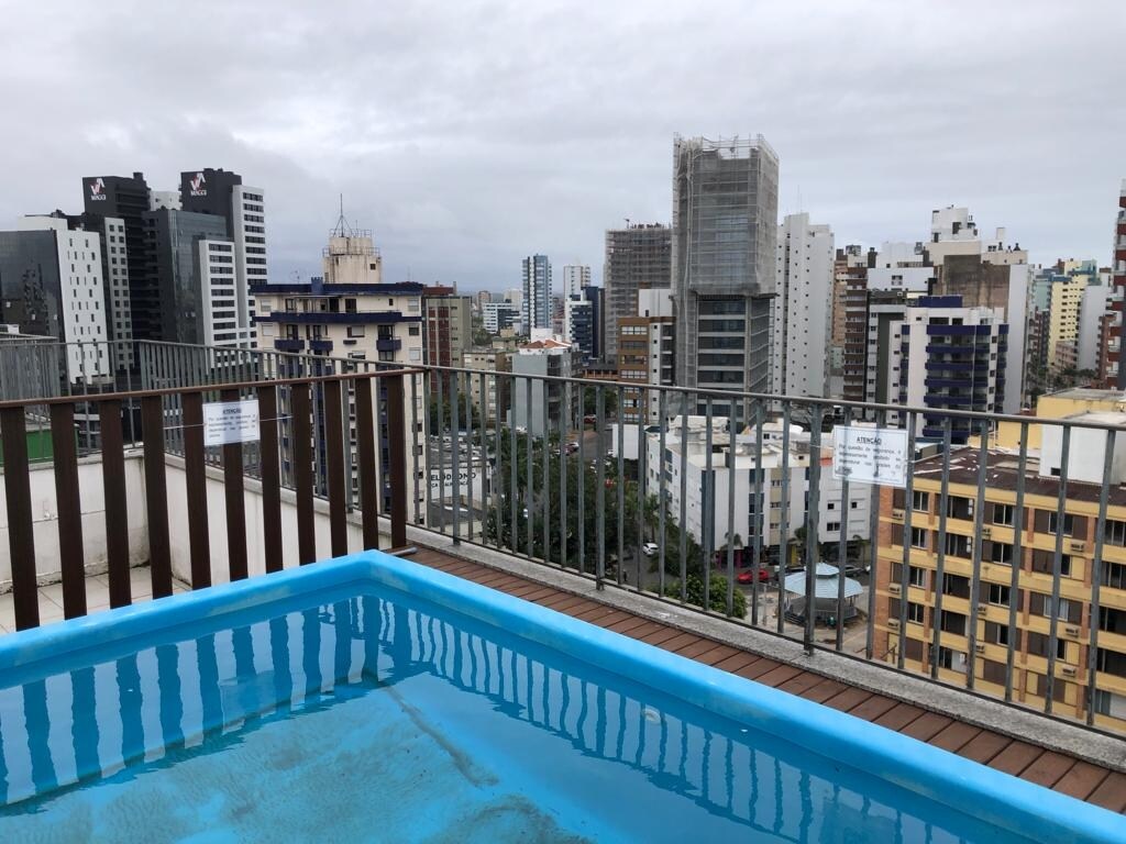A rooftop pool is visible, surrounded by a wooden deck, offering views of the city skyline. Various high-rise buildings and structures are seen in the background, under an overcast sky, creating a serene urban atmosphere.