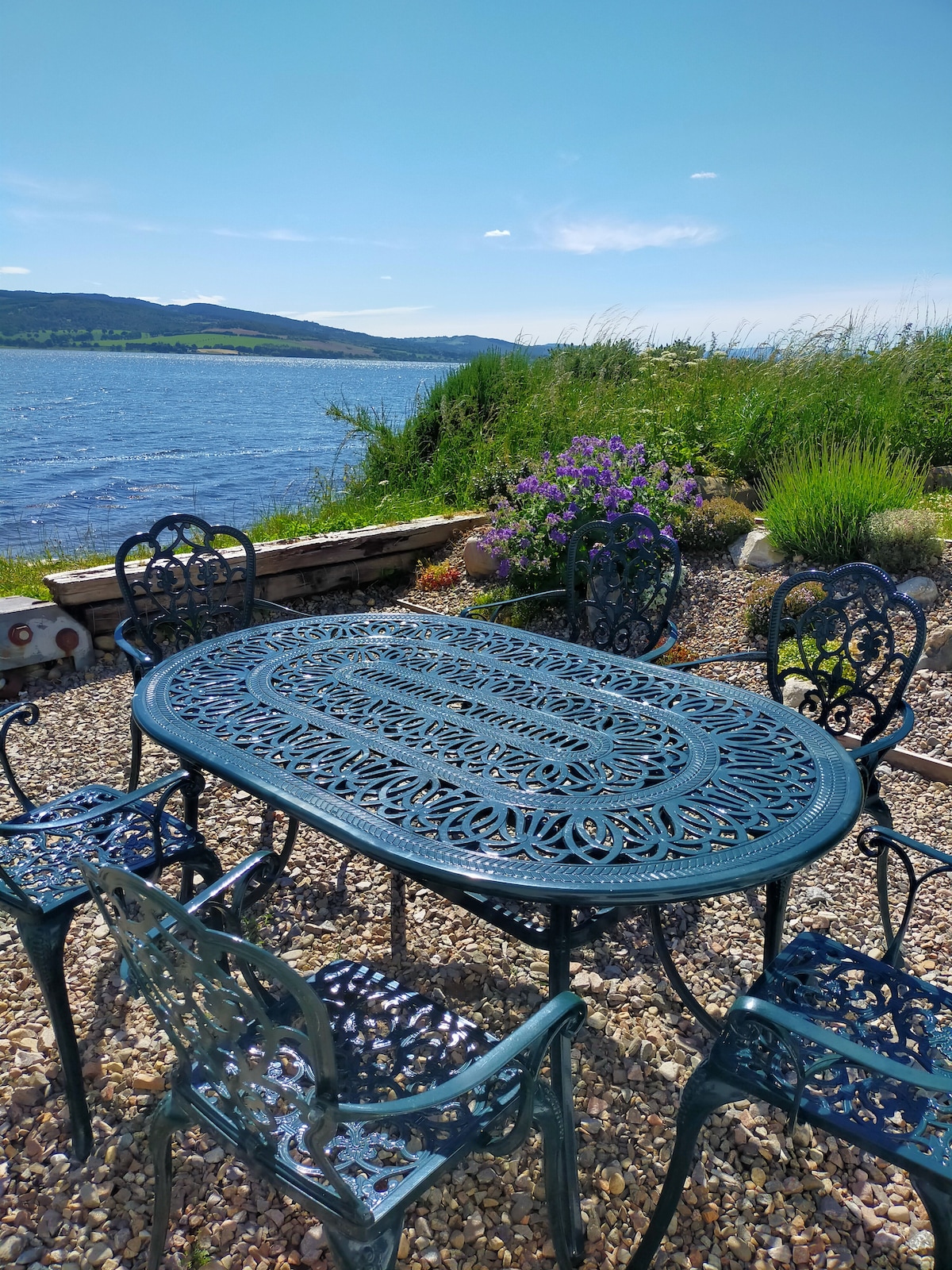 An outdoor setting features a wrought iron table surrounded by six matching chairs, positioned on a gravel area. The backdrop includes views of the Beauly Firth and surrounding greenery, creating a serene environment for dining or relaxation.