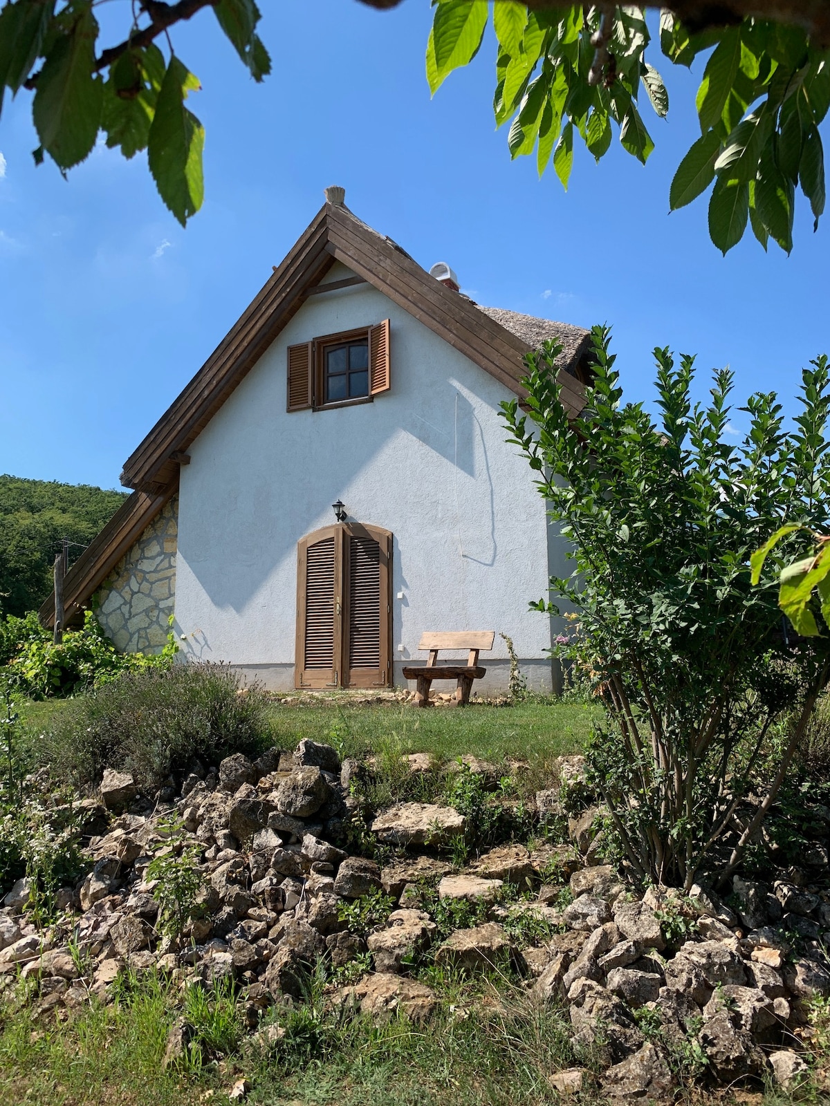 A charming house is set against a clear blue sky, showcasing a sloped roof and wooden shutters. A wooden bench is placed near the entrance, surrounded by lush greenery and rock formations that enhance the natural setting.