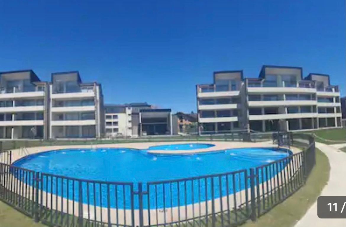 A large outdoor swimming pool is surrounded by modern multi-story buildings, featuring spacious balconies. A separate circular hot tub is visible adjacent to the main pool. A fence encloses the area, with grassy spaces and clear blue skies enhancing the setting.