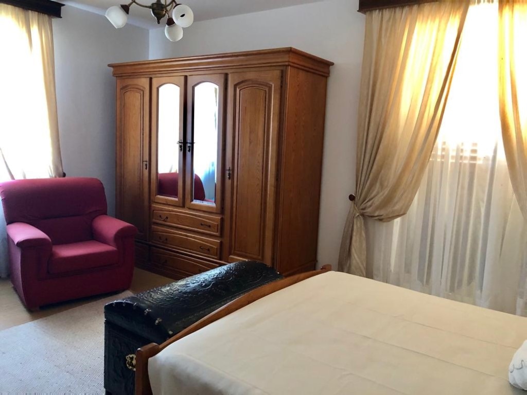 A cozy bedroom features a spacious bed with a light-colored blanket and a wooden chest at the foot. An elegant wooden wardrobe stands against the wall, complemented by a red upholstered chair. Soft curtains and a window allow natural light to fill the room.