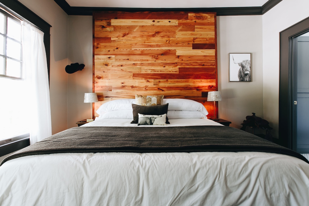 A king bed is centered against a wooden accent wall, with soft lighting enhancing its features. Two bedside lamps are positioned on either side, and decorative pillows add subtle texture. A framed image of a longhorn is displayed on the adjacent wall.