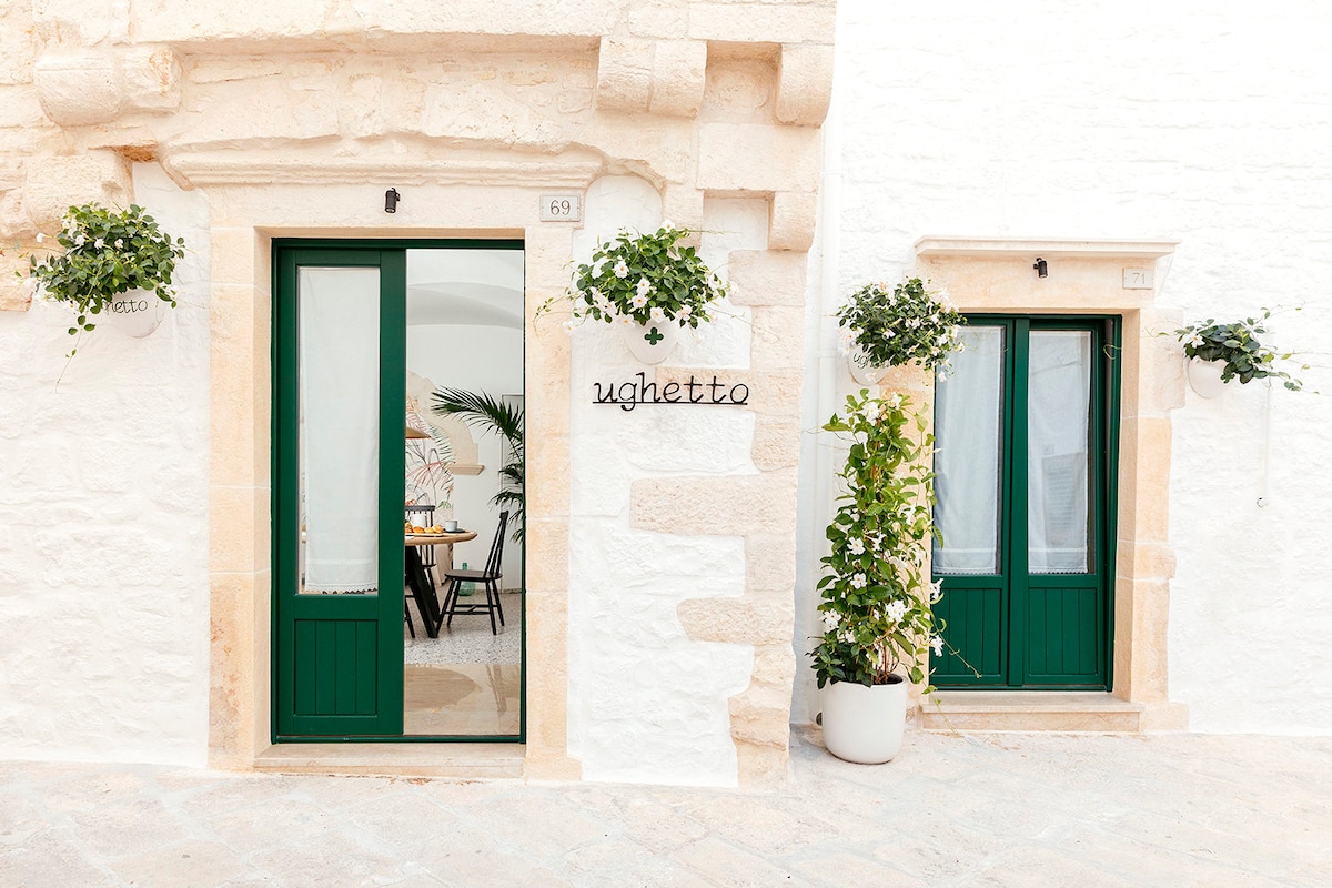 The entrance to Ughetto is highlighted by two green doors framed by light-colored stone walls. Potted plants hang above the doors, adding a touch of greenery. The contemporary signage reading 'Ughetto' is affixed to the wall, offering a welcoming appearance.
