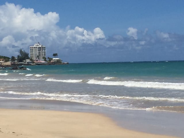 The image captures a serene beach scene with soft waves gently lapping against the shore. A distant building is visible along the coastline under a bright blue sky dotted with fluffy clouds, creating a tranquil atmosphere typical of oceanfront locations.