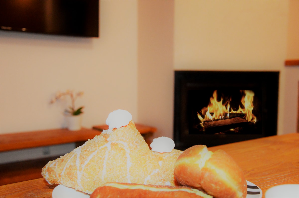 A warm fireplace flickers in the background, adding a cozy ambiance to the scene. In the foreground, a plate of freshly baked pastries is displayed, including a flaky croissant topped with cream and a soft bun, inviting a leisurely breakfast experience.