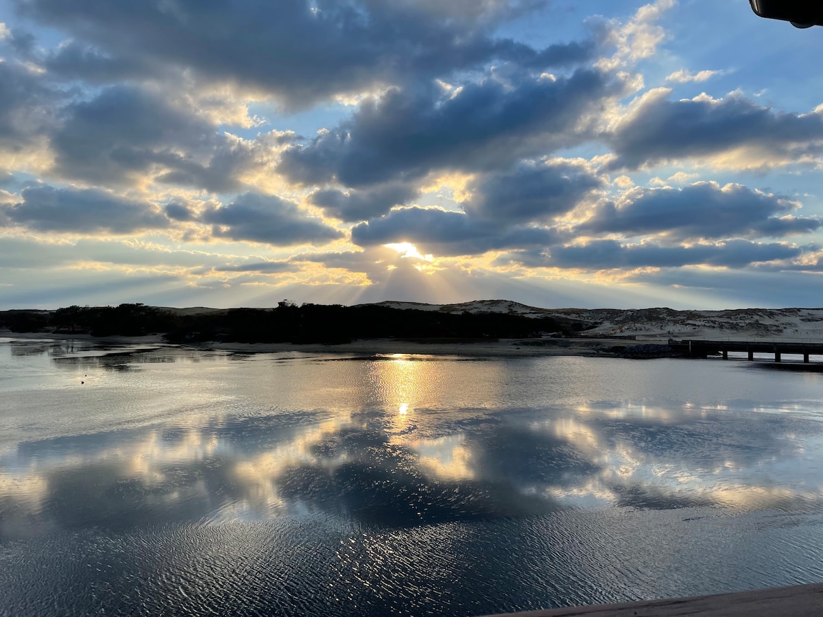 The image captures a serene view of a lake at sunset, with clouds reflecting on the water's surface. The horizon showcases a distant line of dunes, illuminated by the warm hues of the setting sun. Soft waves gently ripple across the lake, creating a calm ambiance.
