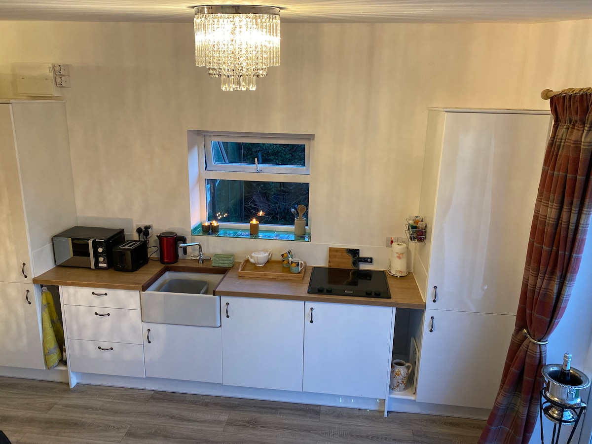 A well-equipped kitchen area features modern appliances including a microwave, kettle, and coffee maker. A large sink sits under a window that offers natural light. Wooden countertops provide ample space for meal preparation, complemented by clean white cabinetry.