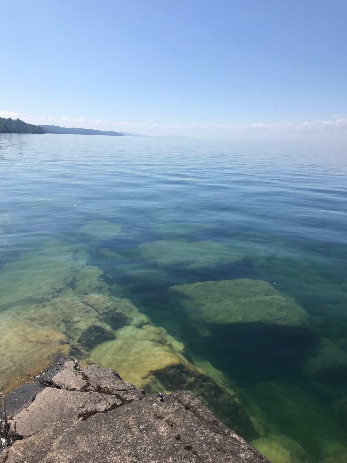 A calm body of water reflects a clear blue sky, creating a serene atmosphere. Beneath the surface, smooth rocks and pebbles are visible, surrounded by gentle ripples. The edges of the shore reveal the gradual transition from land to water, showcasing the area's natural beauty.