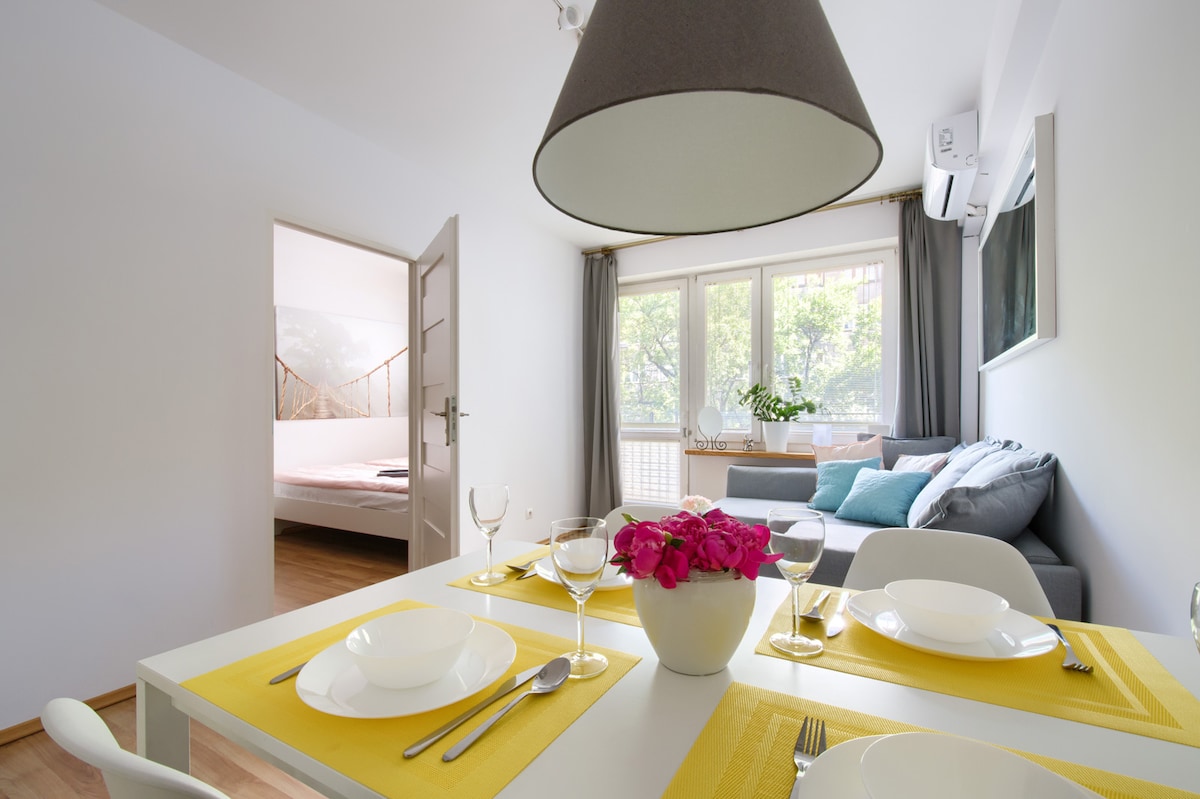 A dining area features a white table set for four with yellow placemats, plates, and glasses. A cozy living room is visible in the background, showcasing a grey sofa and large windows allowing natural light. A vibrant flower arrangement adds a touch of color.