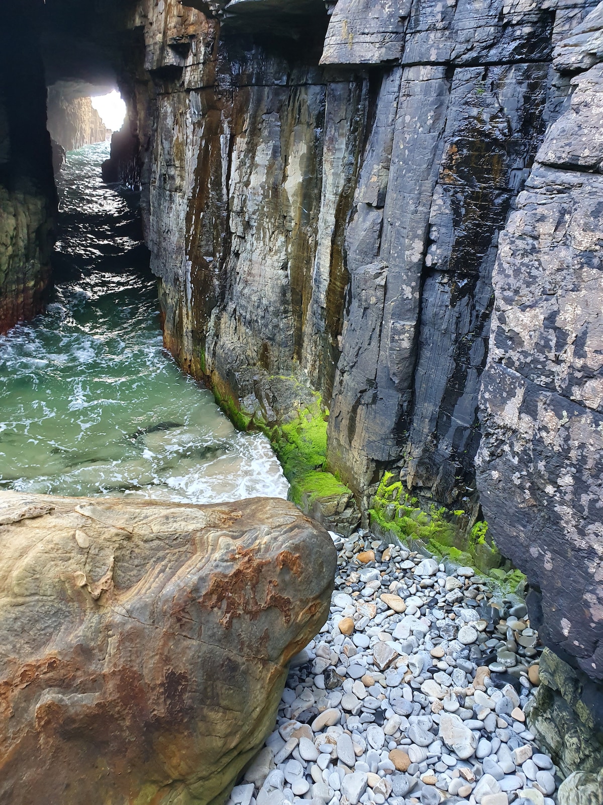 A rocky coastal cove is captured, showcasing towering dark stone cliffs on either side. The water flows gently, reflecting shades of green with patches of seaweed along the edge. Smooth pebbles and larger stones are visible along the shore, adding texture to the landscape.