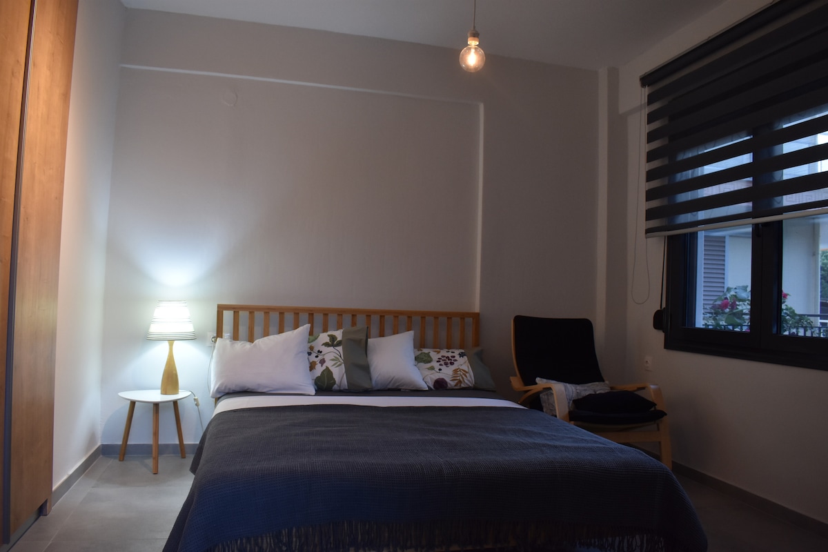 A well-lit sleeping area features a spacious bed with decorative pillows and a cozy blanket. A minimalist nightstand with a lamp offers soft illumination, while a comfortable chair is positioned nearby. Sunlight filters through the modern window coverings, creating a serene ambiance.