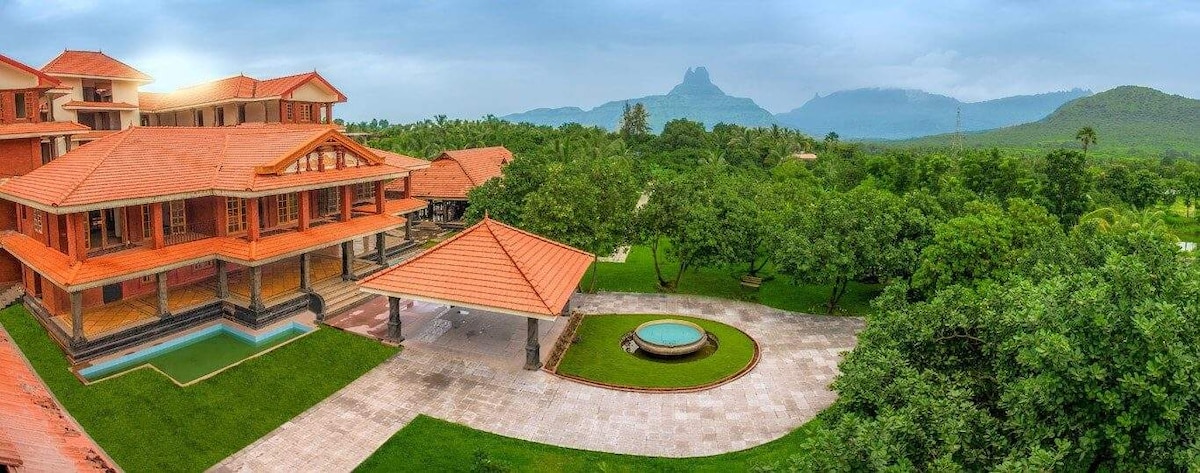 The expansive property showcases a Kerala-style building with a vibrant terracotta roof. Lush greenery surrounds the area, complemented by a circular fountain and a swimming pool. Majestic hills are visible in the background, enhancing the serene atmosphere of the space.