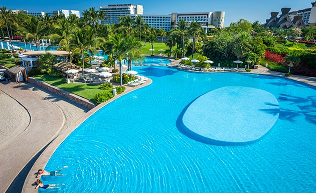 A large, serene pool is surrounded by lush landscaping and palm trees. Sun loungers are positioned nearby for relaxation. The area features a gradual entry point and an island in the center, providing a tranquil setting for guests to enjoy.
