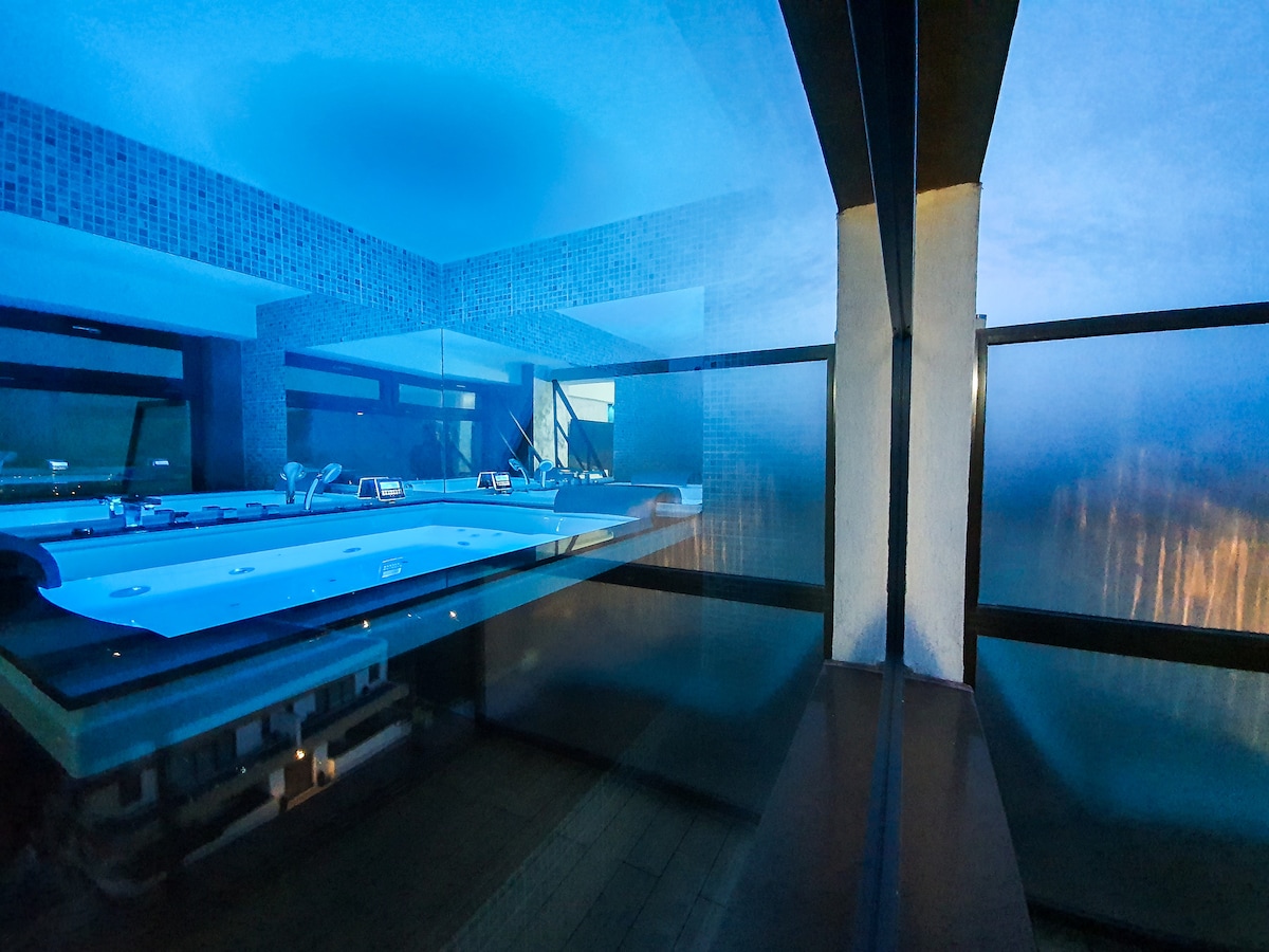 A modern hot tub is positioned near large windows that provide views of the city. Soft blue lighting enhances the ambiance, while reflections create a serene atmosphere. The space features sleek surfaces and minimalist design, contributing to a calm and inviting setting.