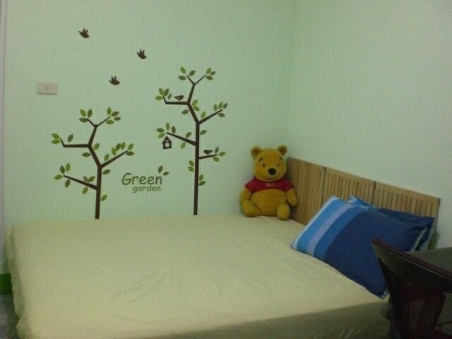 A sleeping area features a spacious bed with a light beige coverlet. A friendly bear plush sits beside blue and white striped pillows against a wall adorned with tree decals, enhancing the room's calming atmosphere.