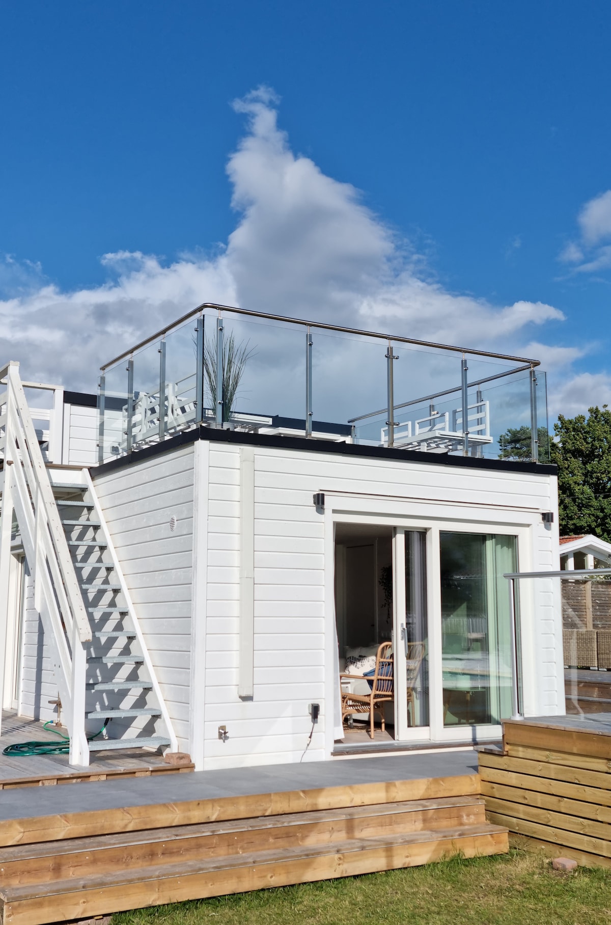 The exterior of the house features a modern design with a spacious rooftop terrace enclosed by glass railing. A staircase leads to the terrace, which is equipped with seating for guests. Large glass doors provide access to the interior from the patio area.