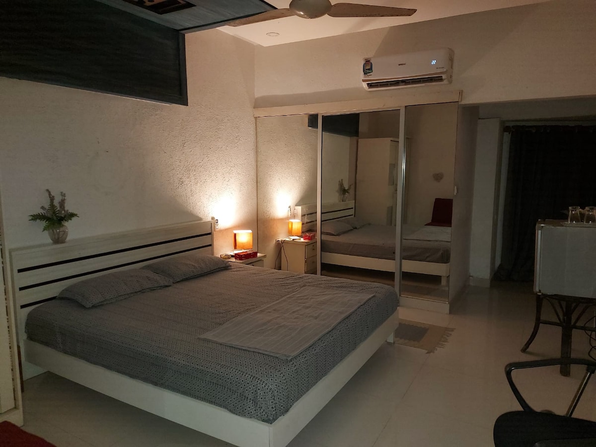 A spacious bedroom features a large bed with neutral bedding. Two bedside lamps provide soft illumination next to mirrored wardrobes, reflecting a simple and functional décor. Air conditioning is mounted on the wall, ensuring a comfortable environment.