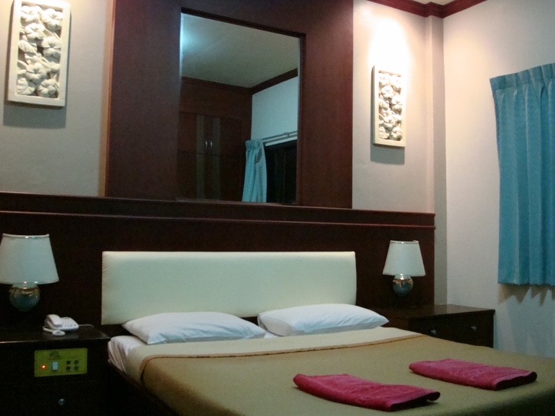 A well-appointed guestroom is equipped with a spacious bed and bedside tables. Two lamps provide soft lighting, and decorative artwork is displayed on the wall. A window with blue curtains allows natural light to enter, enhancing the neutral color palette of the room.