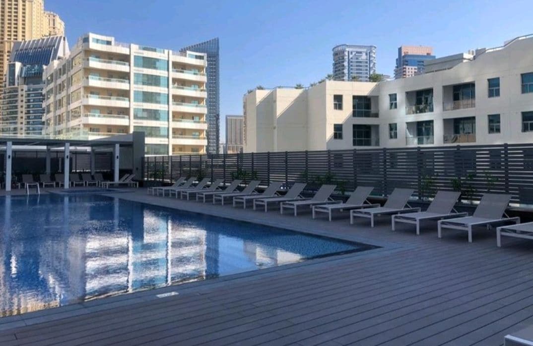 A spacious pool area is displayed, featuring a large, shimmering pool surrounded by a wooden deck. Numerous lounge chairs are arranged neatly along one side, with buildings in the background reflecting a modern architectural style.
