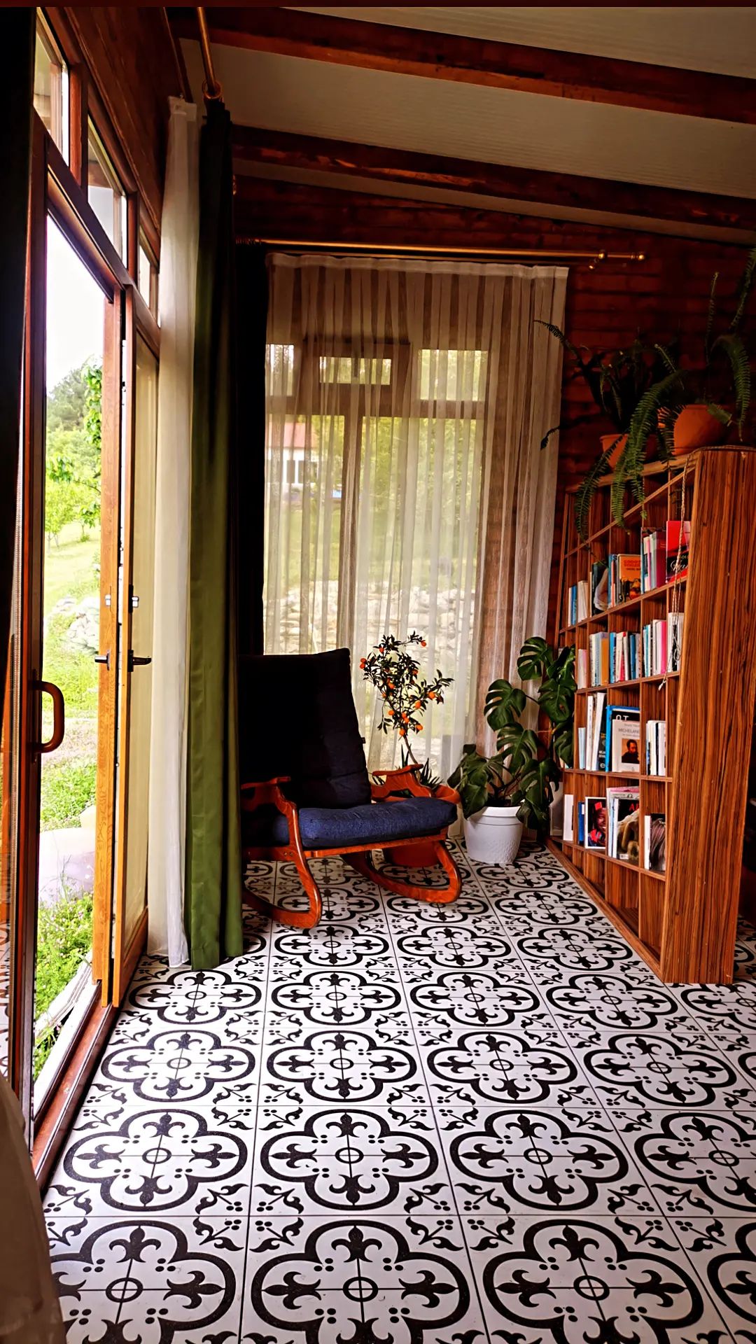 A bright corner of the accommodation features large glass doors, allowing natural light to fill the space. A wooden bookshelf is filled with books, and nearby, a decorative plant adds a touch of greenery. The patterned floor provides a distinctive backdrop for a comfortable chair.