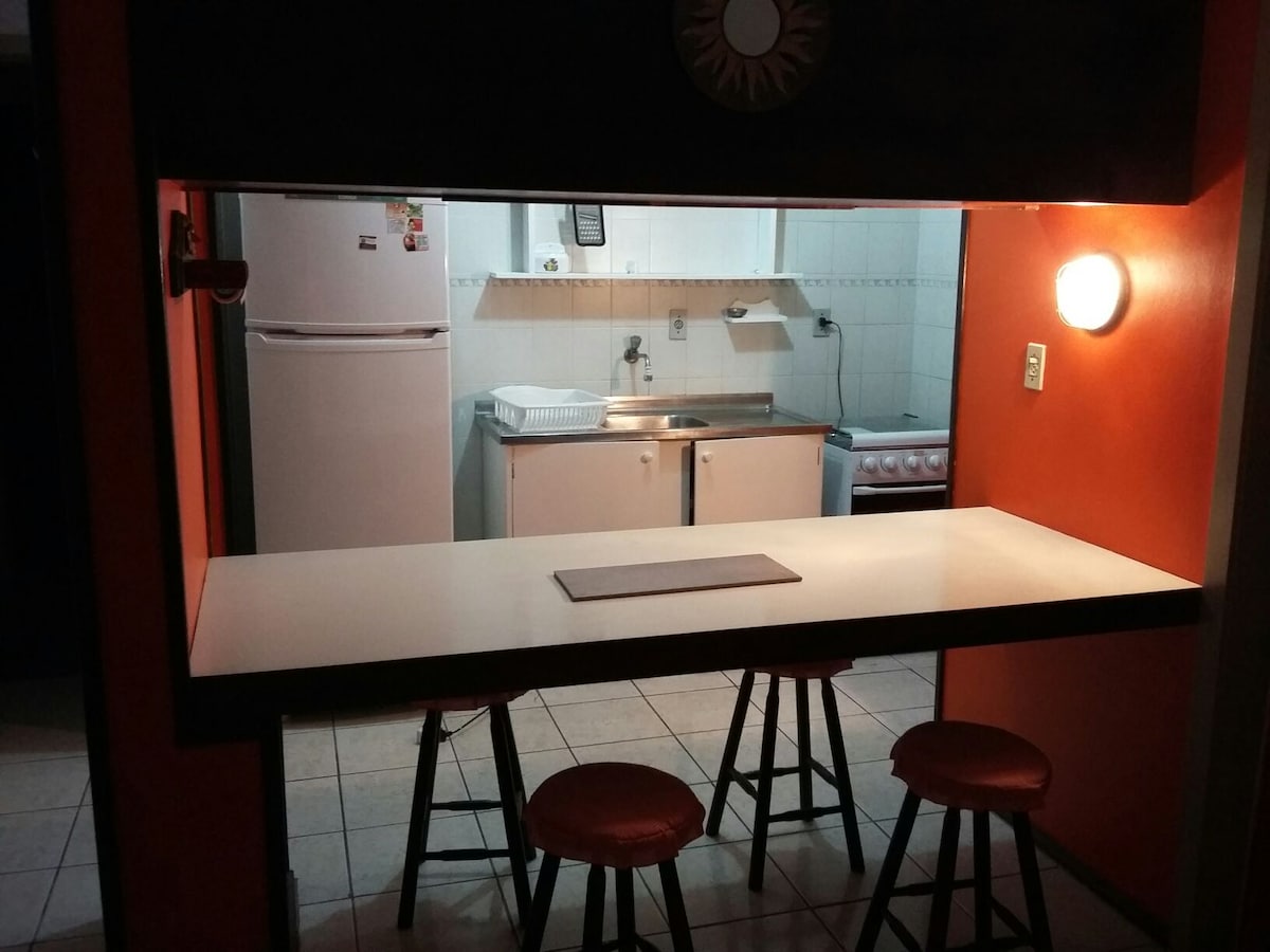 A kitchen area features a well-equipped cooking space with a white refrigerator, sink, and stove set against light-colored cabinetry. A counter with three stools offers a casual dining option, illuminated by a wall-mounted light and complemented by an orange accent wall.