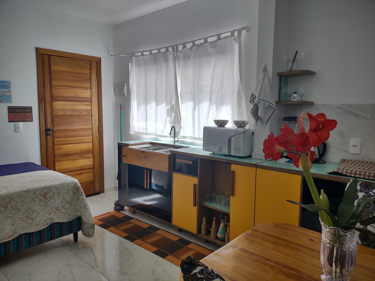 A bright kitchen area features a mix of wooden and yellow cabinetry, with a sink, microwave, and flower arrangement on the counter. Natural light filters through sheer curtains, illuminating a cozy sleeping space with a bed covered in a patterned throw. A wooden door provides entry.
