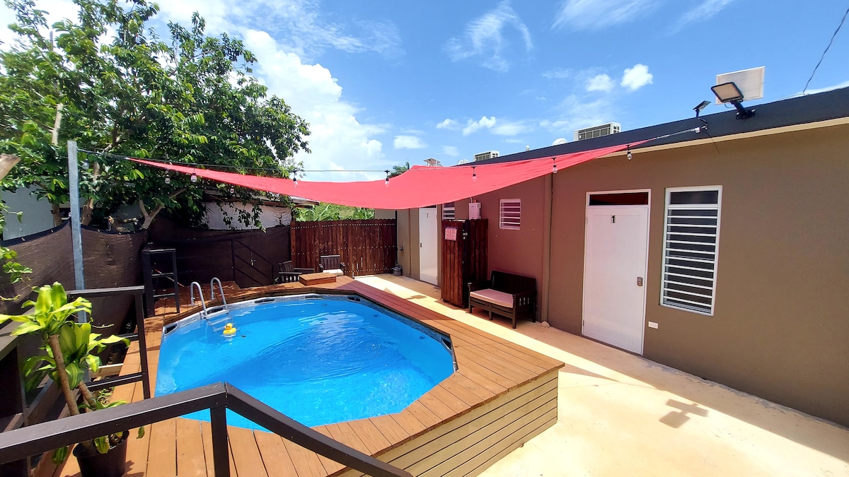 An inviting outdoor space features a sparkling blue pool surrounded by a wooden deck. A shaded area is provided by a red canopy, while potted plants add greenery to the setting. A wooden railing and sunlit walls enhance the relaxed atmosphere.