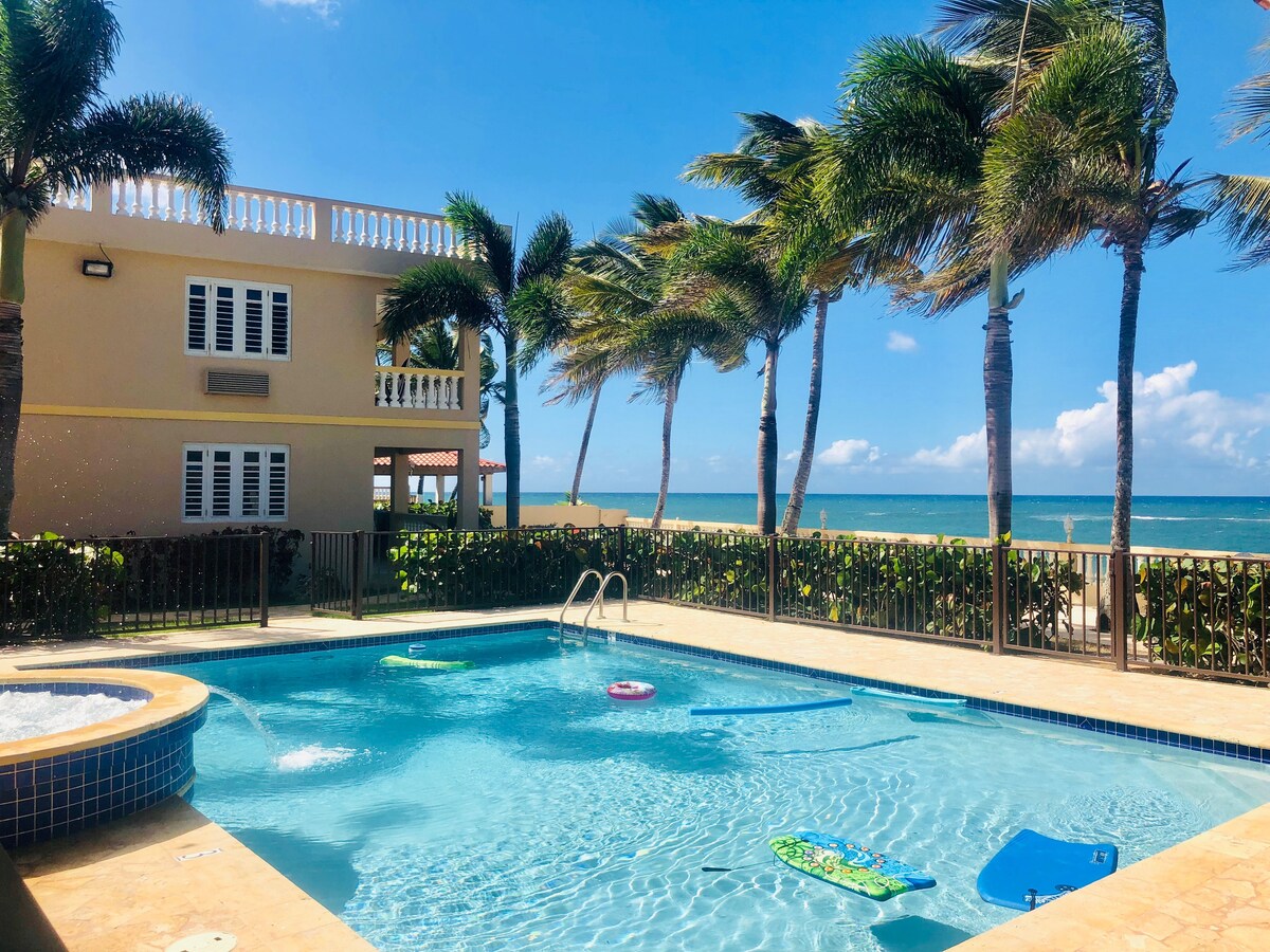 A tranquil pool area is surrounded by palm trees, offering a view of the ocean. The clear water is complemented by colorful pool floaties. The building in the background features multiple windows and a balcony, creating an inviting atmosphere.